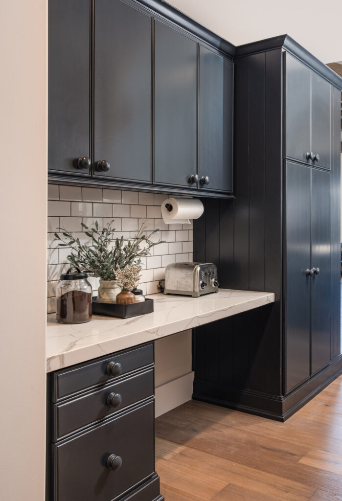 Coffee bar in a luxury kitchen with dark cabinetry, wooden knobs, subway tile with contrast trim. There is a toaster and olive branches styled along the bar.