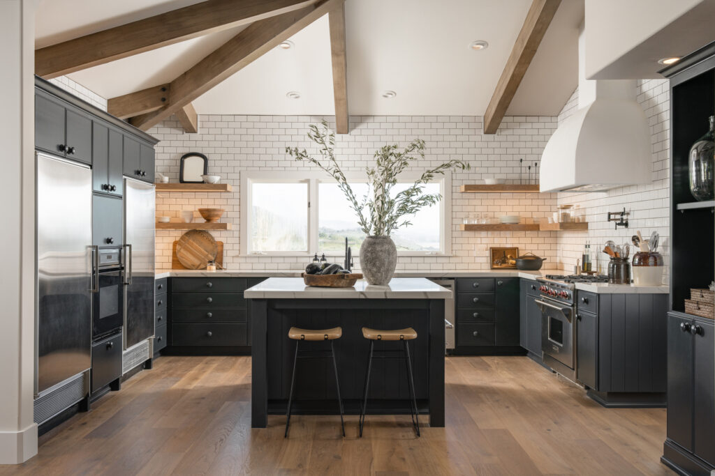 Modern farmhouse kitchen with blue/black cabinetry and painted wooden knobs. Open white oak shelving styled by a professional designer with cutting boards, silver pitcher, glass containers, cups, plates, and other kitchen essentials. Wooden counter stools with iron legs with a metal detail and brass stud on the back adding a small amount of design flare. Large kitchen island marble like quartz countertop, large concrete vase with olive branches, vintage wooden bowl full of eggplants. The wolf range has a custom bell-shaped plaster hood. Subzero fridges.