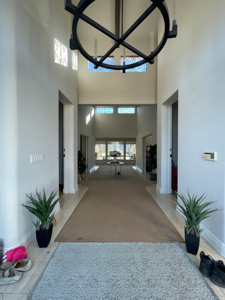 Before picture of an entry of a luxury home located in Santa Ynez in Santa Barbara County California it has carpet a large light fixture high ceilings and large windows displaying beautiful views