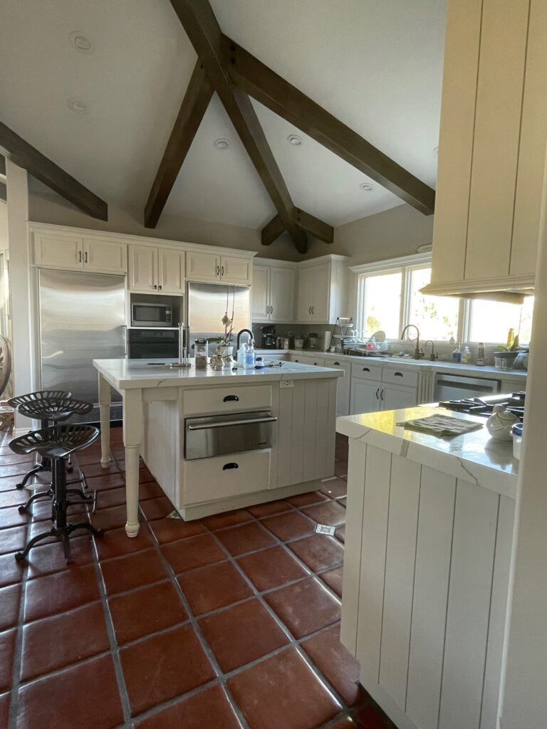 Before photos of a kitchen. Saltillo tile adorns the floor. The traditional style kitchen has white cabinetry with tongue and groove paneling. Large beams finish off the ceiling.