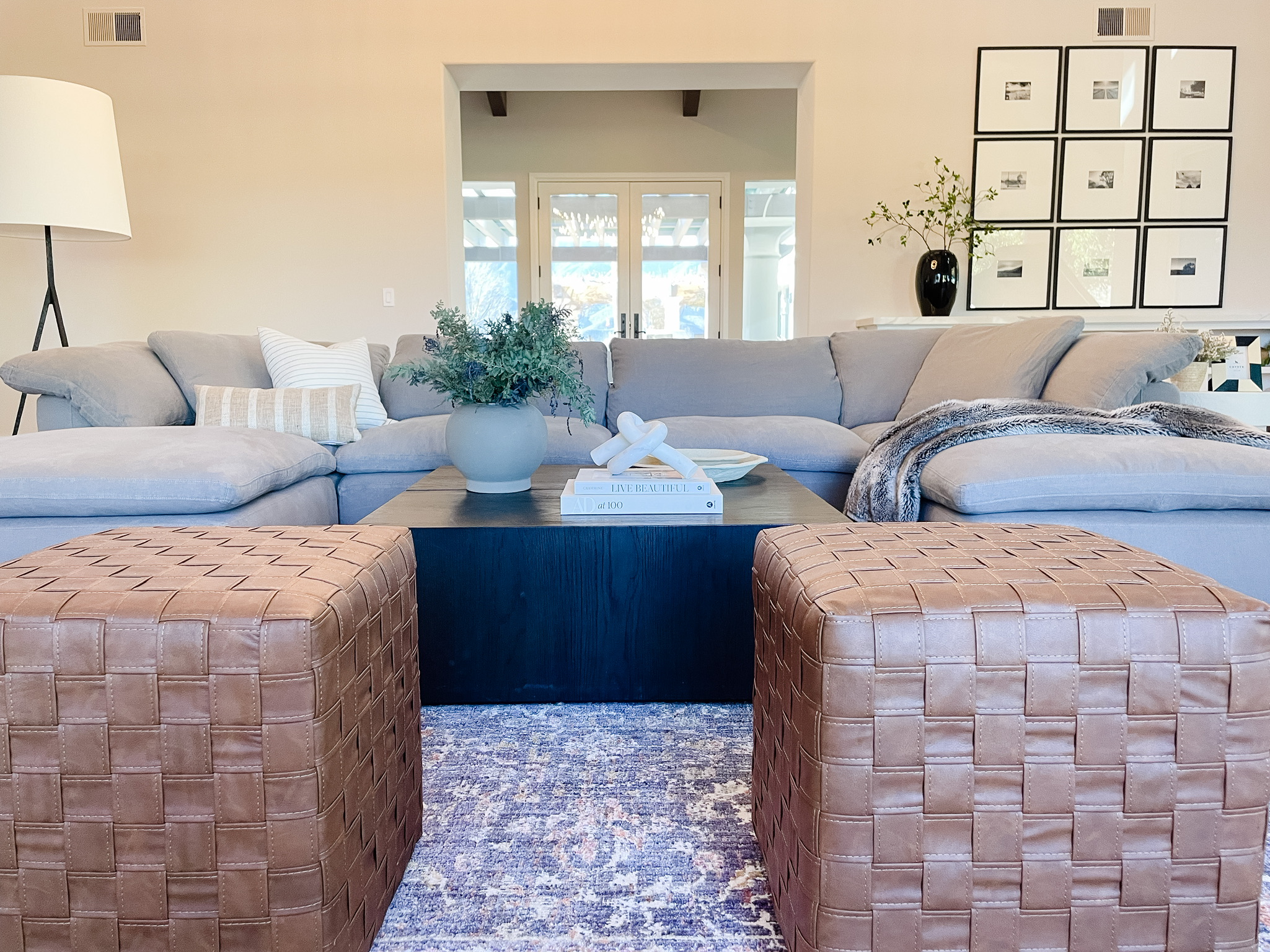 Formal living room of a luxury home that has a large restoration hardware modular sectional in the color fog, two leather woven ottomans, ebonized wood coffee table, chris loves julia x loloi gray Rosemary rug, mcgee and co accessories including a marble knot decorative object, house beautiful coffee table book, AD100, gray vase with green undertone outfitted with green and burgundy branches