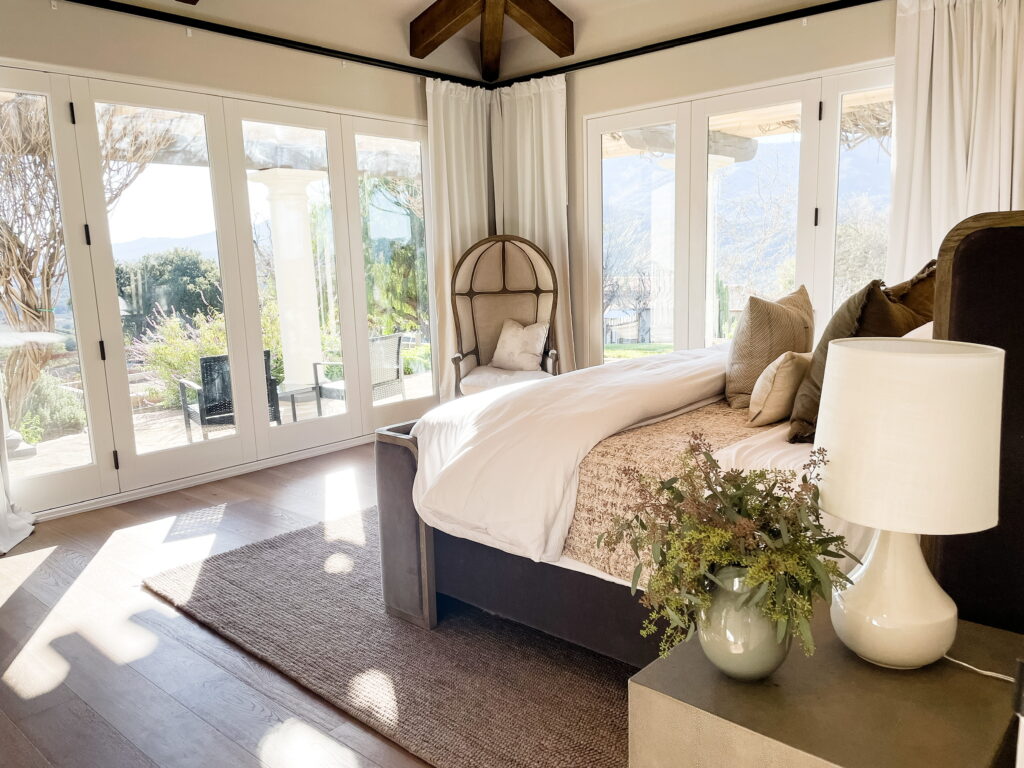 This bedroom in a luxury home has floor to ceiling windows that wrap around the space. There is a white Restoration Hardware Versailles chair in the corner, large cream drapery panels, and a gray velvet Aston bed also from Restoration Hardware. It sit on top a midtone brown hand looped rug and has two Gradon Shagreen Closed nightstands adorned with cream ceramic lamps with white shades that flank the bed. It is finished with a white duvet, brown silk accent pillows, a textural cream lumbar, blue block print pillow, a brown vintage kantha quilt, and a white duvet.