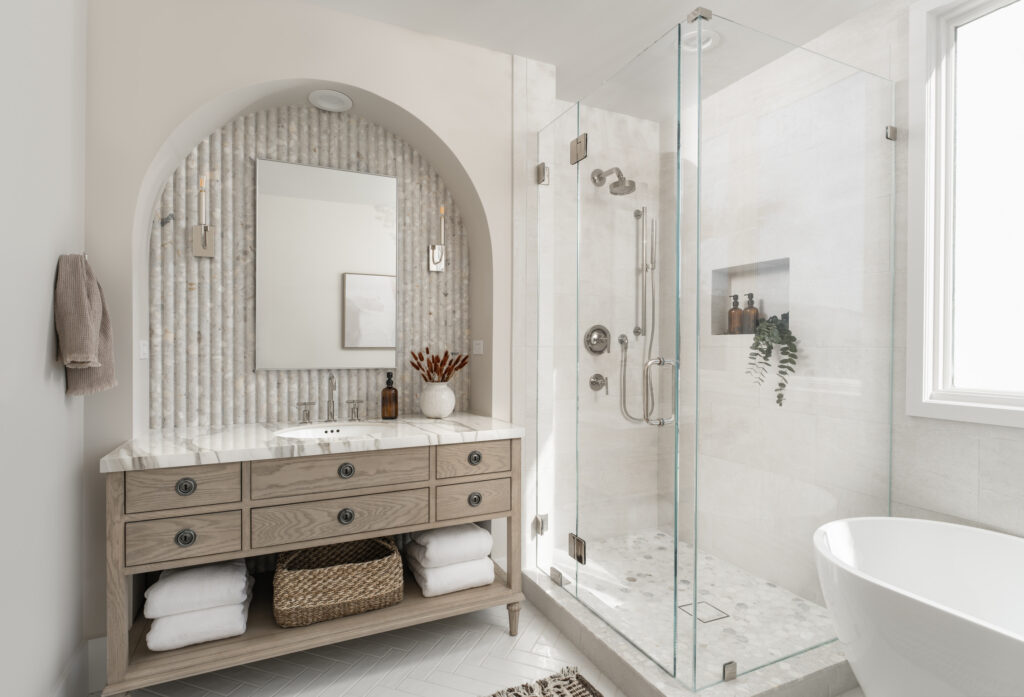 A beautiful arch surrounds a large white oak washstand with drawers and a shelf below. The backsplash is Ann Sacks Terrazzo Renata fluted tile in Brulee. It is a beautiful natural stone with cool grays and warm beige colors against a white back drop. A large frameless shower with glass panels sits next to the vanity with polished nickel plumbing fixtures. the shower has limestone tile that continues along one wall, around the window, and behind the freestanding bathtub. The floor is a matte white subway-style tile in a herringbone pattern.