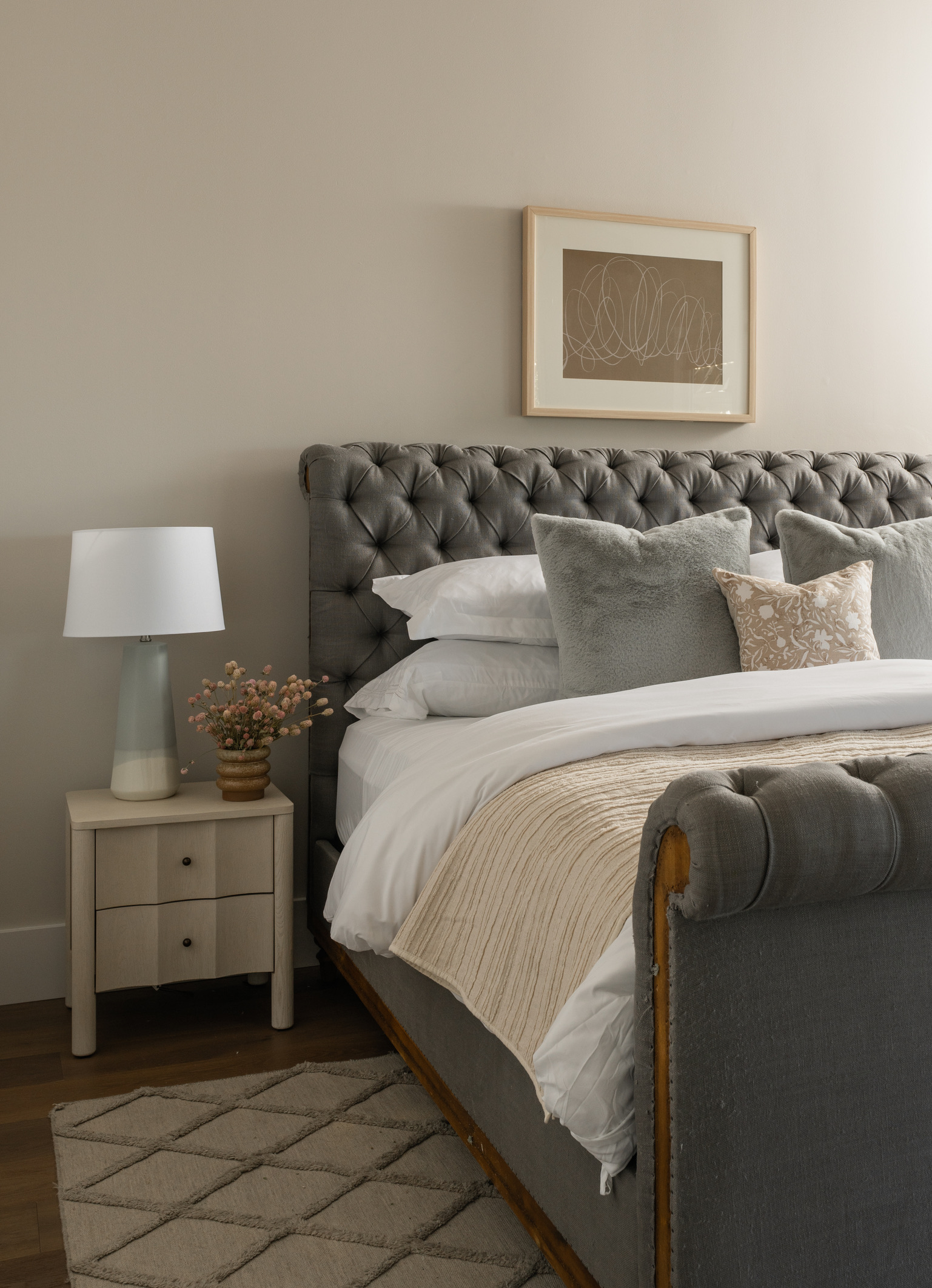 A Napa-inspired black iron chandelier with curved arms sets the tone for this little girl's space. The gray upholstered bed with tufting adds charm to a neutral palette. Medium toned wood beams adorn the ceiling and compliment the similar toned hardwood floors. Blue accent pillows, a light blue and white lamp, and white oak nightstands bring a sense of youth to the space. A taupe and white abstract art piece hangs over the bed in a white oak frame with white matte.