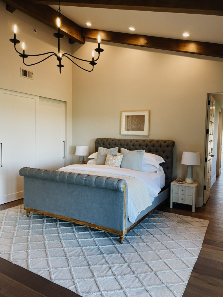 A Napa-inspired black iron chandelier with curved arms sets the tone for this little girl's space. The gray upholstered bed with tufting adds charm to a neutral palette. Medium toned wood beams adorn the ceiling and compliment the similar toned hardwood floors. Blue accent pillows, a light blue and white lamp, and white oak nightstands bring a sense of youth to the space. A taupe and white abstract art piece hangs over the bed in a white oak frame with white matte.